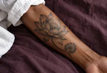 Tatouage fleur de lotus : signification, couleurs et compositions Lotus noir et gris, tige courbe et feuille ronde sur un avant-bras à peau brune.